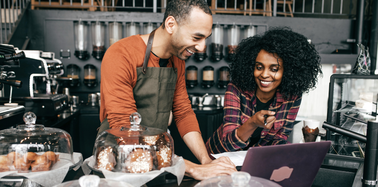 A man and a woman in a coffee shop are standing behind the counter. They are leaning on the counter and looking at a laptop.